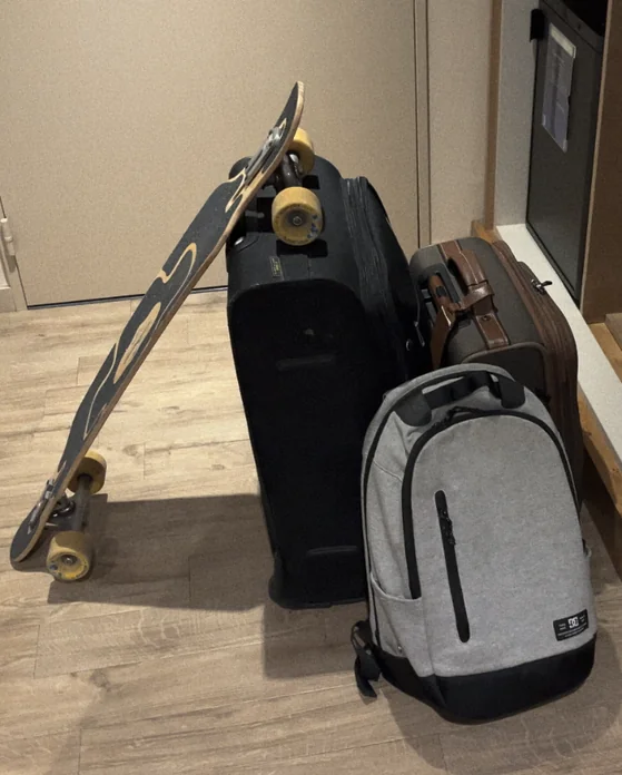 A suitcase, a carry-on and a backpack standing by the door. A longboard is leaning on the suitcase.