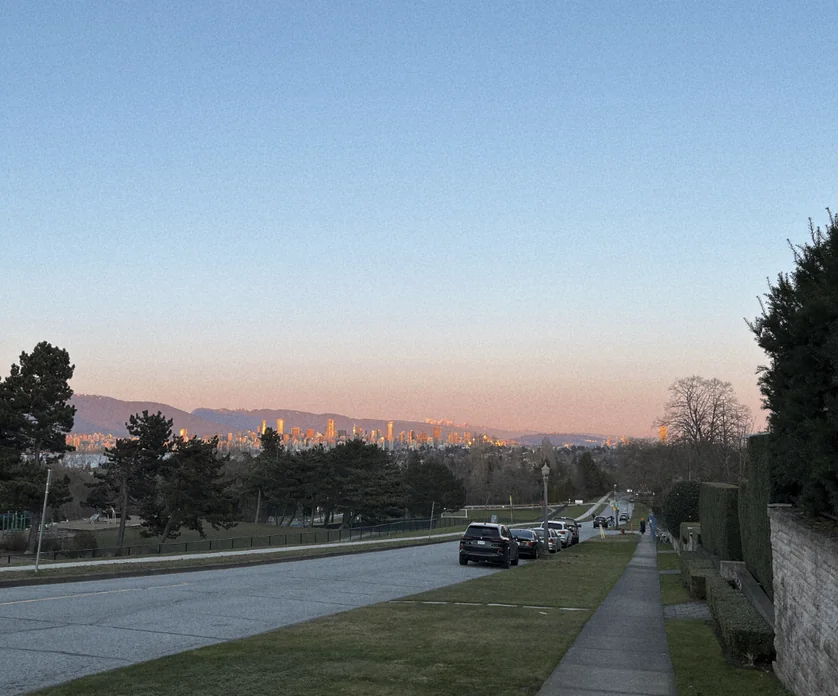 View towards Downtown Vancouver from a hill in West Point Grey at sunset: between nearby trees and faraway mountains, skyscrapers glow red in the Sun.